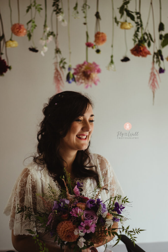 A bride smiles happily under blooms hanging from an archway. The bride wears a lace dress and holds a colourful bouquet/ the individual large blooms are very colourful. A hint of the brides wheelchair can be seen. 