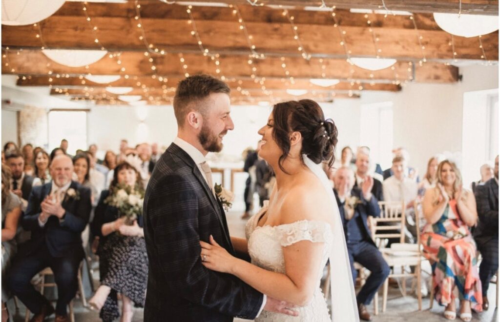A wedding couple stand facing each other smiling. behind them are their friends and family sat either side of the asile. The bride wears a white off the shoulder gown and the groom a subtly checked black suit. The room is white, light and bright with oaks ceiling beams.