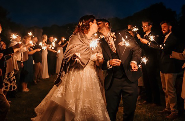 A wedding couple kiss at the end of a sparkler parade. Guests stand either side holding sparklers. It is night and so dark but the sparkers give off a soft pink glow. The bride wears a lace white dress and is wrapped in a shawl. 