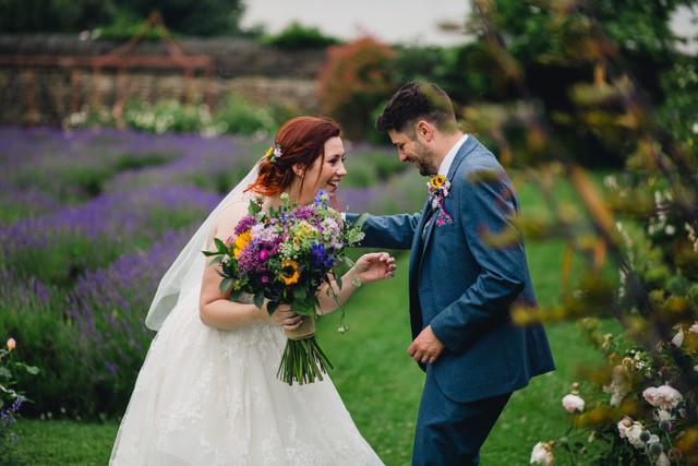 A bride and groom laugh together. They are stood slightly aprt but reaching for one another. She wears a whit gown and holding a brightly colored floral bouquet. He wears a blue suit. They stand in front of a purple lavender labyrinth, Tudor stone walls with white roses to their right. 