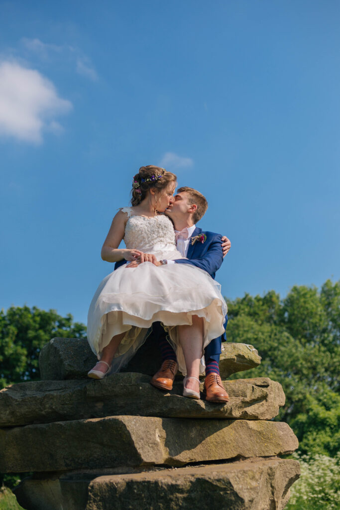 A wedding couple sit on top of a stack of large rocks. The bride sits on the grooms knee. the bride wears a short lace dress and the groom a blue suit. They are kissing. The sky is pure blue and there are green trees in the background. 