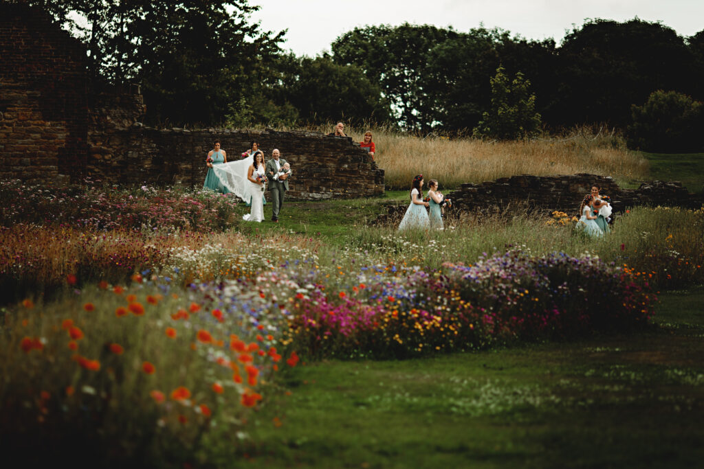 A bride in a  white gown walks with her father, in a  grey suit, down an outside wedding aisle filled with white, pink, purple and red flowers. She is preceded by two adult bridesmaid is pale green dresses and a flower girl in a white dress. Another bridesmaid holds her train. In the back ground are an old stone wall and trees. 