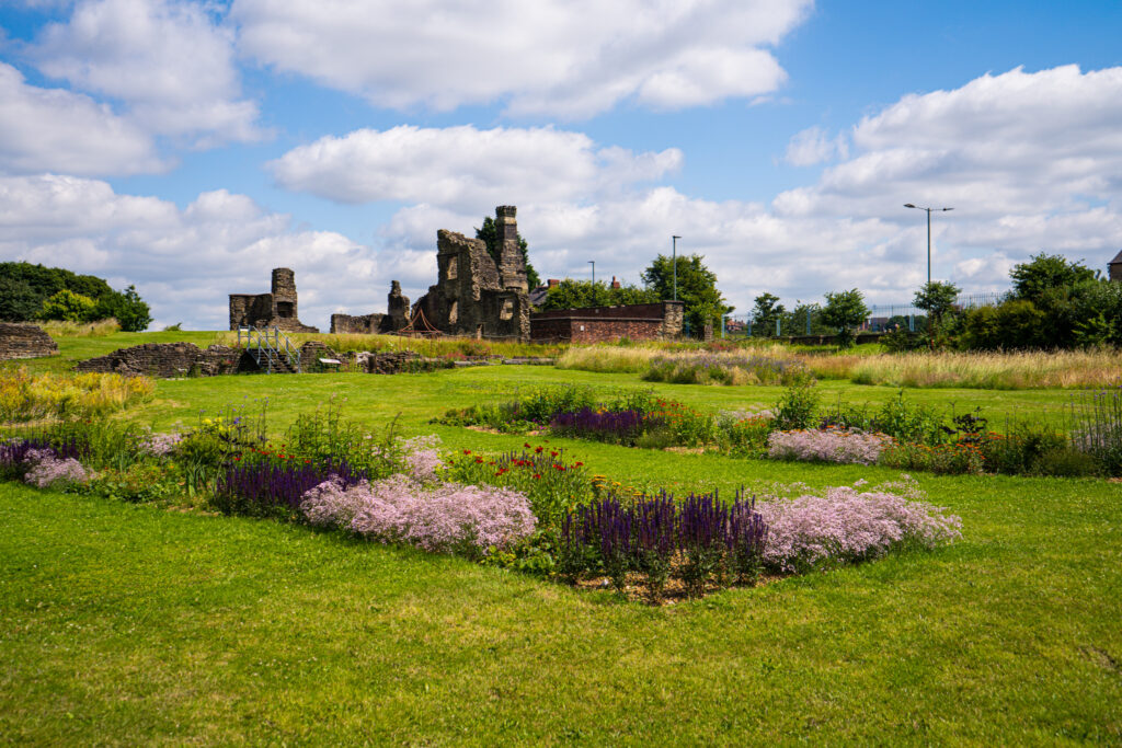 Tudor stone ruins stand in the middle ground of the photo. They are ruinous but two chimneys, a fireplace and a wall with three windows can be seen. In the foreground is a grassed area covered in perennial planting. There are dark and light purple flowers including baby breath and salvias as well as hints of red. In the background is long grass and perennial meadows. the sky is blue with white fluffy clouds.