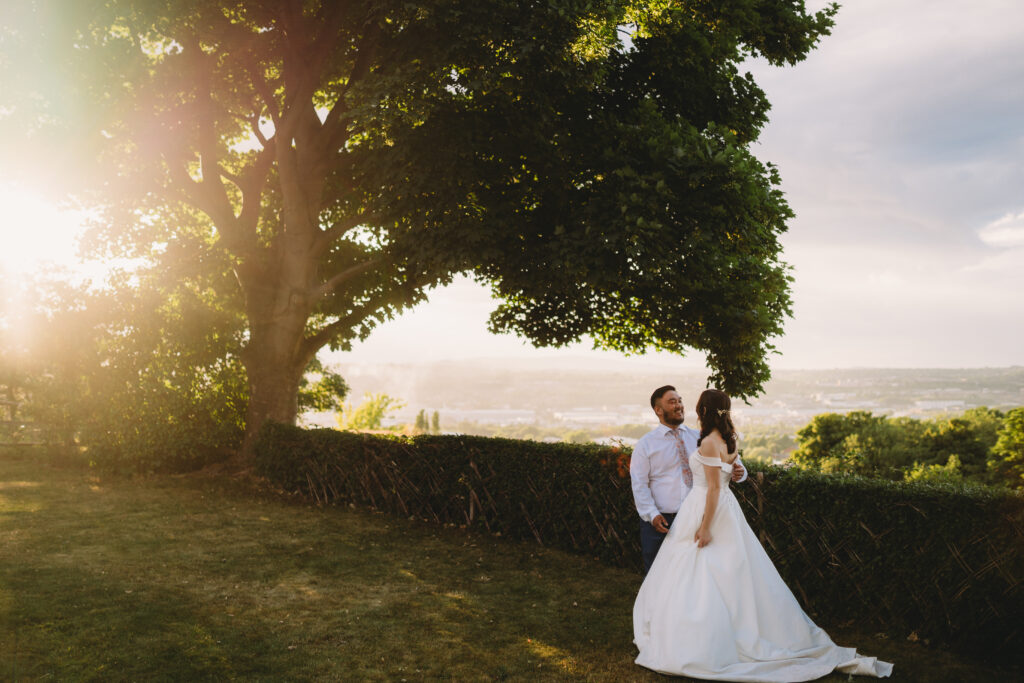 A wedding couple laugh together as the sunsets. They stand close. She wears a full white wedding dress. He wears a suit with jacket removed. They stand on grass over looking a viewpoint of trees and hills. The light blurs the background. To their right is a large oak tree with the sun shining through. 