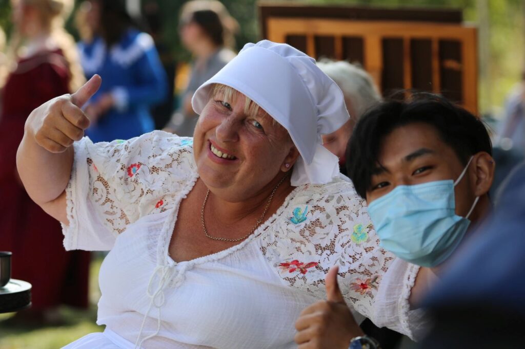 A volunteer dressed as a Tudor maid smiles happily at the camera with her thumbs up. She is with a younger male volunteer who is wearing a facemask.