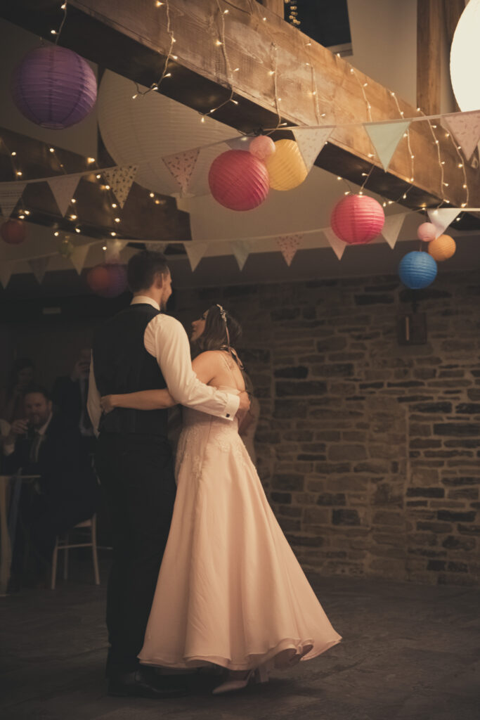 A wedding couple dance under oak beam covered in fairy lights and colourful paper lanterns. The room is dim but their is glowe from the subtle lighting. Bunting covers the ceiling. The bride wears a flowy pink dress and the groom a traditional black suit. They have their arms around each other as they dance. 
