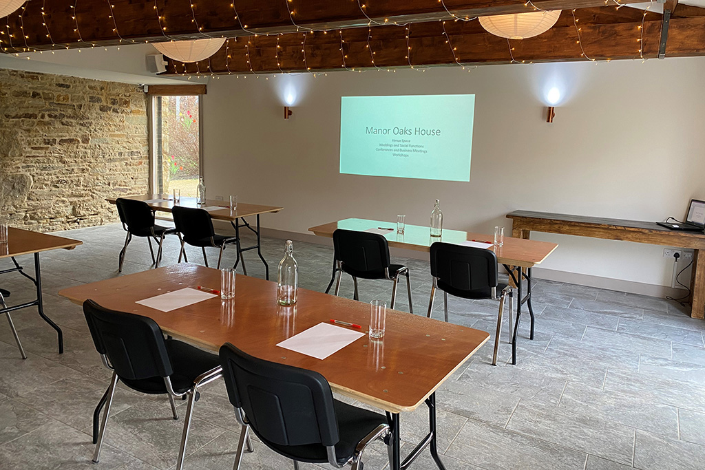 A bright light room topped with oak beams and a slate grey floor. There are four wooden desks each with two black conference chairs facing a projector screen. On the table are water bottles, glasses, pens and paper. A wooden table sits in the corner with a laptop on it.
