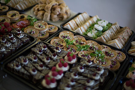 A close up of a buffet spread that includes cake, mini quiches and sandwiches. the sandwiches are carefully cut into circles and triangle. The quiches are garnished with green herbs and sprinkled with feta cheese. The cakes are in the foreground and include chocolate cake cut into triangles with a dark cherry on top and cupcakes covered with raspberries.