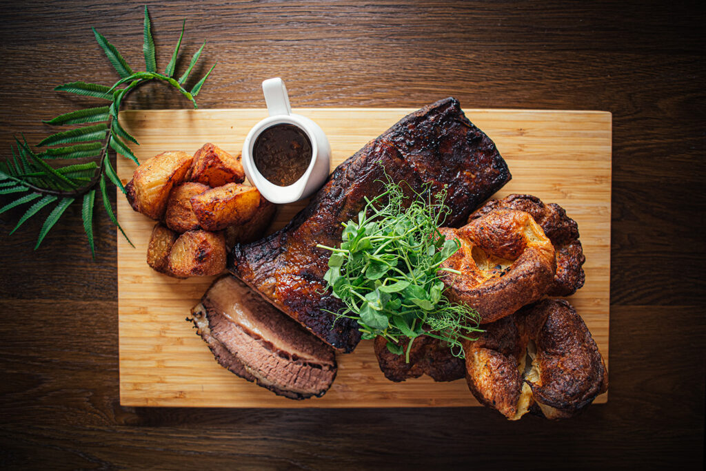 A glazed roast beef joint sits on a wooden serving tray. Two slices have been cut at the end and laid flat. it is cooked medium. Next to the joint sit three Yorkshire puddings and a stack of roast potato's . The plate is garnished with green salad and there is a whit just with gravy. The serving board wits on a dark wood tables with a fern leaf for decoration.