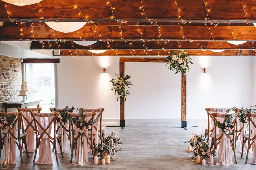 A wooden arch is covered in large bouquets of pink flowers. It stands at the end of an aisle of wooden chairs covered in pink flowers and draped with pink tulle. The room is white and light. On the ceiling are oaks wooden beams covered in fairy lights. 