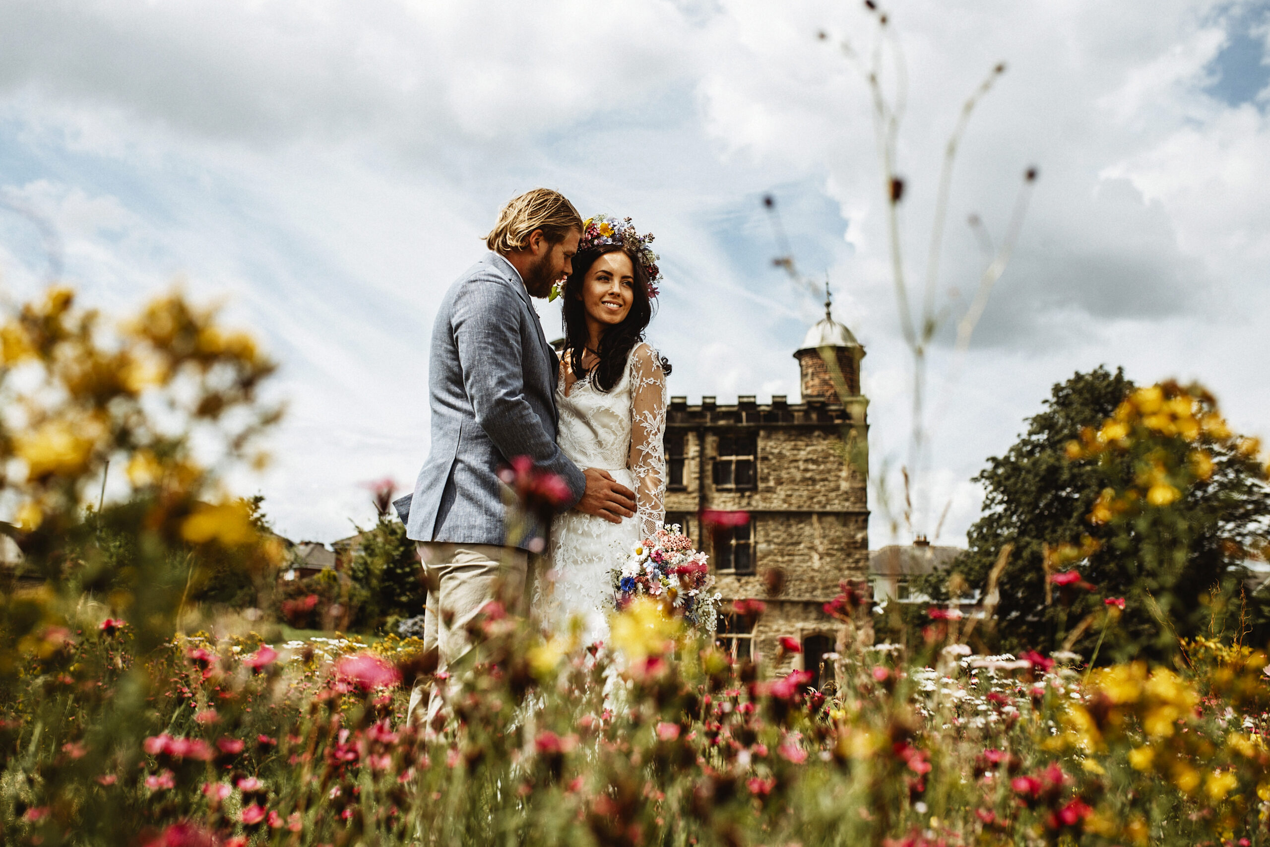 A young wedding couple stand in front of the stone Tudor Turret House at Sheffield Manor Lodge. The bride wears a lace white dress and a flower crown. The groom wears a light blue suit. He holds her as she gazes towards the camera. In the foreground are yellow and pink meadow flowers.