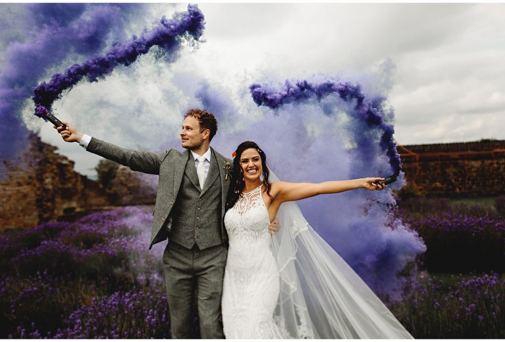 A wedding couple stand next to each other, one arm around the other, the other arm outstretched holding a purple smoke bomb. The two smoke bombs rise up into the air. They stand in front of a lavender labyrinth, the purple smoke blending with the blooms. 
