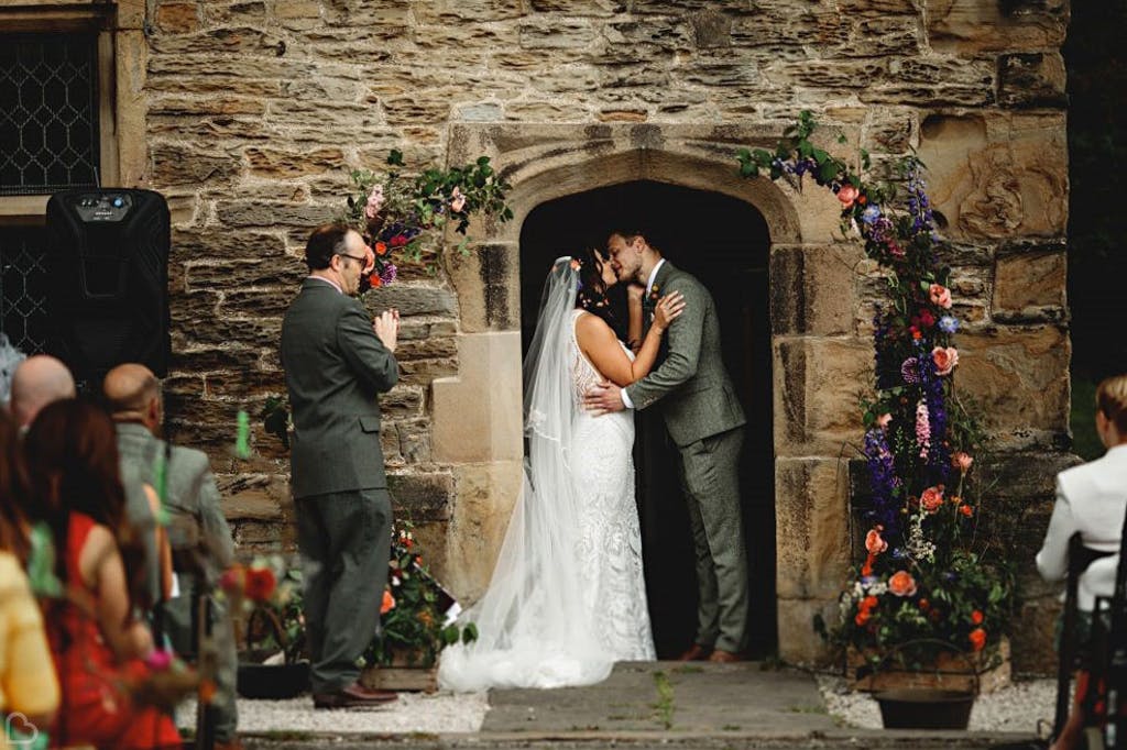 A wedding couple kiss in the doorway of a stone Tudor building at the end of their wedding ceremony. The bride wears a white lace dress and the groom a grey suit. Pink and purple floral arch surrounds the door. Guests sit facing them clapping. 