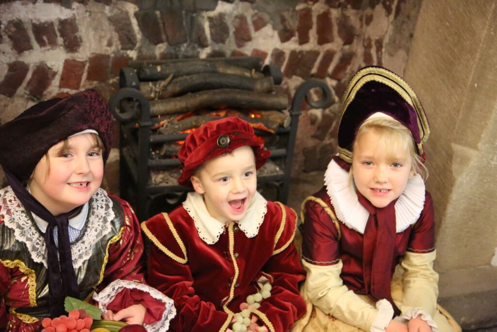 Three children aged 4 - 7 sit in a Tudor stone fireplace. There is a grate in the background which glows red with red light that represents fire. The girl on the left smiles while looking slightly off camera. She wears purple velvet gable hood and red silky dress with a lace ruff. Her school uniform peeks underneath. The both in the middle is open mouthed with delight. He wears a red velvet cap and matching tunic with gold trim. The tunic has a lace collar. The girl on the right wears a purple and gold gable hood while smiling directly at the camera. He dress is red silk and gold with a large white ruff.
